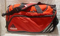Front view of red Adidas flight bag with black trim and Adidas logo on side pocket, showing general shape and features.