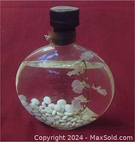 Front view of clear round glass bottle filled with jasmine-scented bath oil, containing white beads and a white artificial flower inside, with black cap on top, sitting on a red surface.
