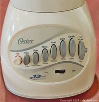 Close-up of Oster blender base showing 12-speed control buttons and function labels (Pulse, Grate, Blend, Shred, Grind, Liquefy, Whip, Ice Crush, Mix).