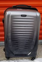 Front view of a metallic grey hard case suitcase with horizontal ridges and a central Samsonite logo badge on the front panel.