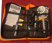 Photo of the whole soldering iron kit inside black and orange zippered case showing soldering iron power cord, metal stand, solder spool, tips secured in yellow backing plastic, and soldering flux bag.