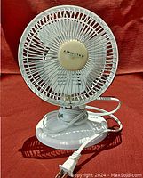Side angled photo of white Airworks desk fan showing grille, blades, base, and attached power cord with plug, set against a reddish background.