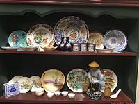 Display of multiple large decorative plates, two blue candle holders, blue and white porcelain box, small bowls, and a spouted white ceramic vessel on shelves.
