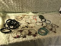 Photo shows a collection of various bracelets laid out on a textured cloth. Included are multiple gold and silver-tone bangles, black cord bracelets, and turquoise stretch bracelets.