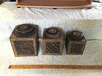 Three square wood canisters with metal inlay and floral lids, arranged largest to smallest alongside a ruler for scale.