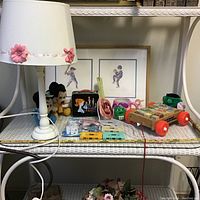 Shelf containing white lamp with flower details, framed children artwork, Mickey Mouse plush, character mugs, metal lunchbox, toy cars, and wooden push toy.