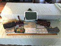 Photo showing a group of 11 evening bags, one floral fabric book holder with handle, and one wooden yardstick, arranged on a beige blanket on a couch.