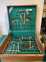 Flatware set in open wooden case with green velvet lining showing knives, forks, spoons, and serving utensils in designated slots.