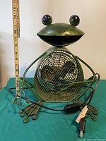 Front view of green metal frog shaped fan showing wire cage, frog eyes, arms and power cord with switch coiled in front, measuring about 14 inches tall.