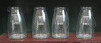 Photo of four identical clear glass lanterns lined up side by side, showing their cylindrical shape and metal handles.