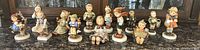 Full lot of 16 Hummel porcelain figurines displayed on a surface showing all figures.