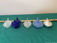 Front side view of 5 mini chicken-shaped glass covered dishes in different colors on green cloth with ruler for scale