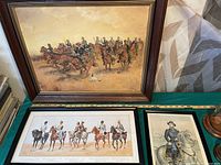 Framed military paintings depicting cavalry and mounted soldiers arranged on green fabric surface.