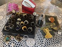 Top-down photo showing multiple costume rings on a black display foam, wristwatch, two Artifacts pins in packaging, a pink beaded pouch, a silver-tone rhinestone hair comb in a box, and a black ring sizing tool.