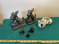 Photo of vintage Scottie dog silver bookends, ceramic cream pitcher, ceramic ashtray, and three small Scottie dog figurines/keychains on green surface with measuring tape