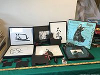 Multiple Scottie dog art and decorative pieces arranged on a teal cloth with various frames and a wooden plaque.