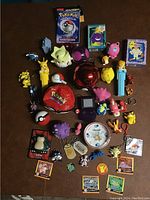 Top down photo displaying a diverse collection of vintage Pokémon toys and collectibles including PEZ dispensers, figures, keychains, a Game Boy, Pokémon card packs, and a clock.