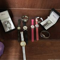 Photo of eight wrist watches displayed on a wooden surface, including two Carolee watches in boxes and six loose watches with varied strap materials and colors.