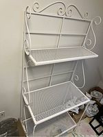 Full side angle view of white metal bakers rack showing three shelves and scroll metalwork.