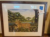 Full view of framed lithograph depicting a big cat (spotted) in a lush natural scene with colorful flowers and trees under glass in a dark wooden frame.