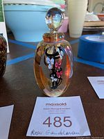 Front view of the signed Roger Gandelman perfume bottle with floral design and stopper sitting on a table next to a MaxSold auction card.