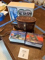 View of four binocular items showing three original boxes (Kamakura, Vanguard, Bushnell) and one brown leather case for the antique German binoculars with shoulder strap.