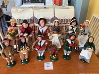 Full view of 12 Thanksgiving and Pilgrim-themed figurines arranged on wooden table with house-shaped boxes behind them.