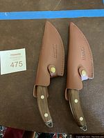 Two Huusk branded cutting tools with wooden handles and metal reinforcements, partially inside brown leather sheaths, shown from above on a dark surface.