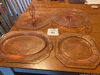 Four pink Depression Glass items on wooden table: a rectangular platter, a large round platter with handle, a smaller round serving plate, and a cake server.
