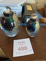 Pair of signed Robert Eickholt hand blown glass perfume bottles shown upright with stoppers attached on a wooden table with a maxsold auction tag.