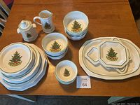 Full view of the Pfaltzgraff Christmas dishes set arranged on a wooden table showing plates, bowls, sugar bowl, creamer, serving platter, and cookie plates with Christmas tree and village motifs.