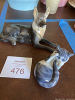 Photo showing a lying German Shepherd dog porcelain figurine with gray and beige glaze alongside a Royal Copenhagen cat figurine in a playful pose with gray and white colors.