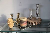 Five assorted candle holders and two candles arranged on a glass shelf with a white wall background, showing overall lot contents and composition.