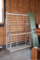 Full view of white metal baker's rack with laminate wood shelf in middle, scrollwork design visible on sides and top.