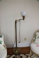 Full view of the brass-coloured floor lamp placed between two chairs against a wall, showing its height and general condition with some visible metal wear.