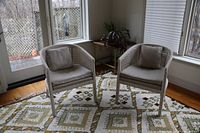 Two white wooden cane back chairs with upholstered seats placed side by side in a bright room with a patterned rug.