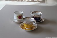Photo showing all three fine china teacups with saucers, two with ornate gold trim and colored bases, one white with rose design.