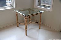 Square wicker side table with glass top placed near window, viewed from an angle showing the corner and one side of the table, highlighting wicker frame and glass edges.