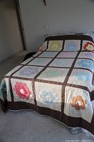 Full view of the handmade quilt laid on a bed; shows colorful floral hexagon patch blocks separated by brown grid lines.
