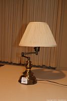 Full view of brass swing arm desk lamp with pleated fabric shade illuminated from the side.