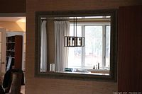 Front view of large bevelled glass wall mirror mounted on wall reflecting window, curtain, and light fixture.