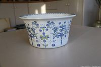 Side view of Lotte hand painted Norwegian casserole without lid showing blue and green floral and human figure artwork around exterior and blue patterned rim.
