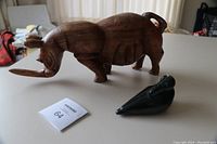 Photo showing both sculptures: a carved teak rhinoceros with a repaired tail and a dark stone bird sculpture.