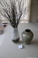 Two large vases on a table, one white-gray with branches inside and one green speckled bulbous vase.