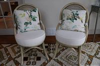 Pair of vintage cane chairs with white round seat cushions and floral pillows, placed on patterned rug near glass-paned door.