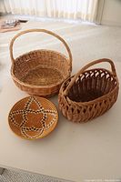 4 wicker baskets, including one round with handle, one oval with decorative weave and handle, and two smaller baskets of different shapes and styles, plus a set of woven placemats featuring a circular star pattern with beads