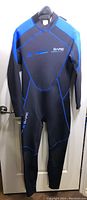 Front view of men's wetsuit hanging on hanger showing black color with blue panels and seams, Bare logo on chest and left arm.