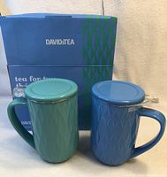 Photo of two Nordic style ceramic mugs in teal green and blue, each with matching lids, in front of David's Tea box.