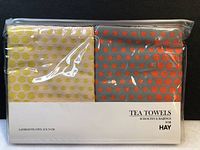 Two folded tea towels in original sealed plastic packaging, showing layered dots pattern in different colors and product label with brand and dimensions