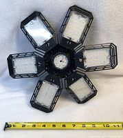 Top view showing six foldable LED panels arranged in a circular, flower-like design with a measuring tape for scale, approx 11 inches diameter.